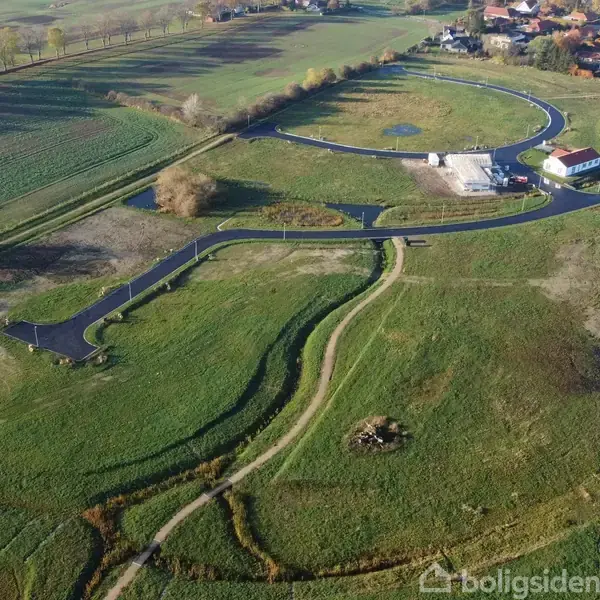Luftfoto over en grøn mark, hvor nyanlagte asfaltveje indrammer et område nær en lille bygning. Området omgives af åbne marker og en bebyggelse i baggrunden.