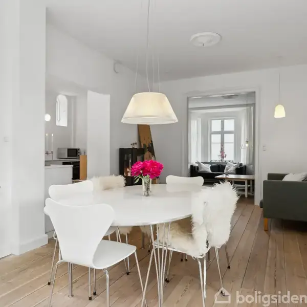 En hvid spisebord med fem stole står i et lyst rum. En lampe hænger over bordet. I baggrunden ses en stue med gråt møbel og flere vinduer.