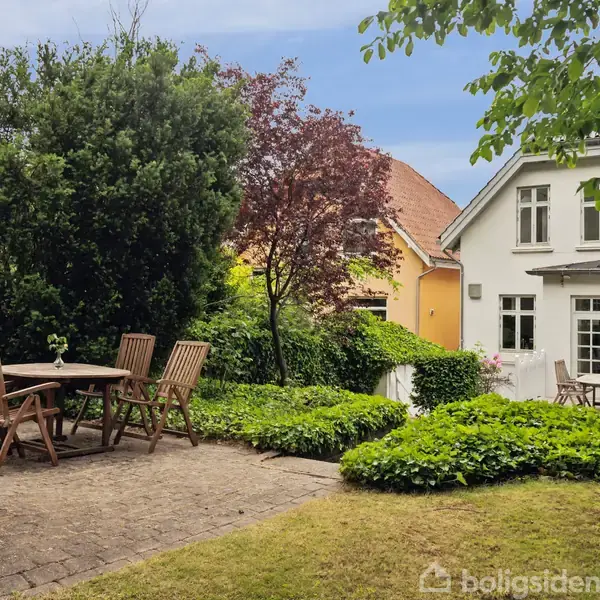 En terrasse med træbord og seks stole står på fliser, omringet af frodig have. I baggrunden ses et hvidt hus med store vinduer og en gul bygning ved siden af.