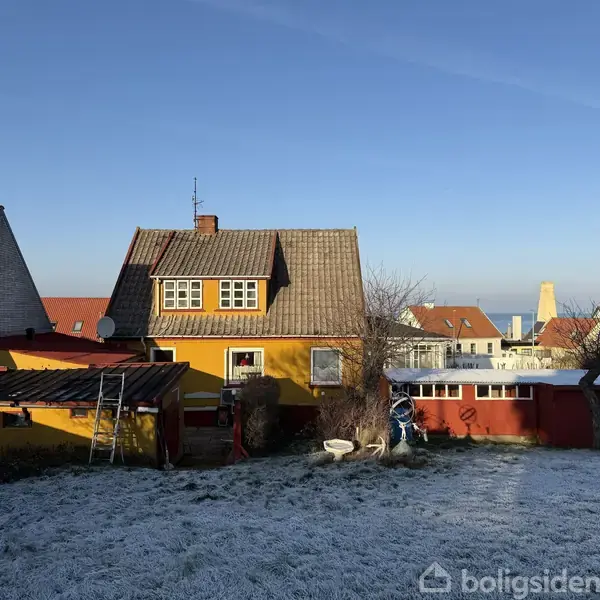 Et gulmalet hus står stille i en frostdækket have blandt flere boliger. Baggrunden viser en klar, blå himmel. En skorsten og antenne er synlige på taget.