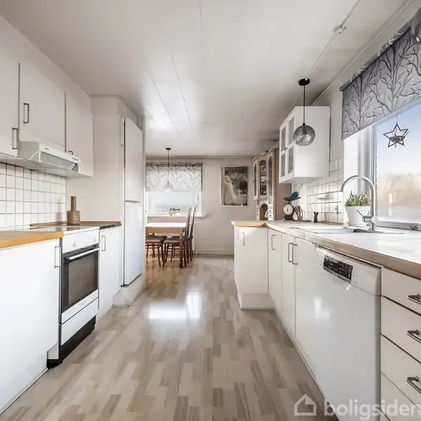 Køkken med hvide skabe og træbordplader står i et lyst rum. En spiseplads med bord og stole ses i baggrunden, omgivet af store vinduer og trægulv.