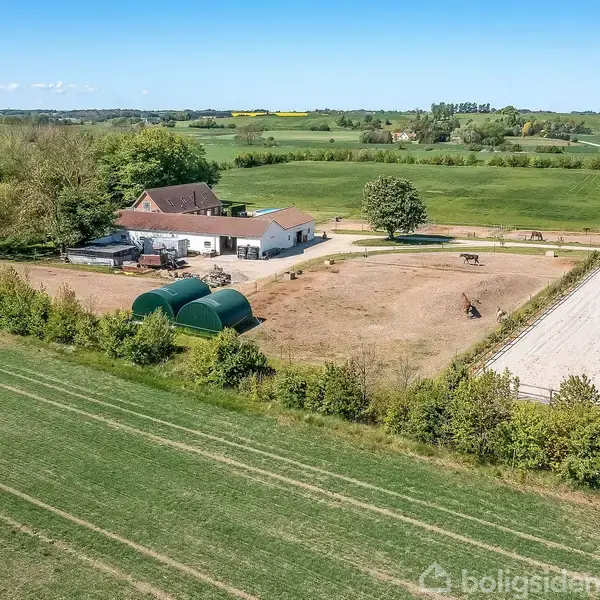 En gård med et hovedhus, omgivet af grønne marker og en indhegning, hvor der ses en hest. Flere opbevaringstelte står tæt på bygningen. Landskabet er frodigt og åbent.