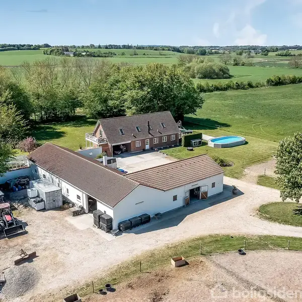 To landbygninger og en swimmingpool er omringet af frodige marker og træer. En grusvej fører til ejendommen, der ligger i et åbent, landligt landskab.