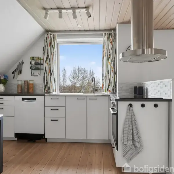 Køkken med hvide skabe står stille. Et bord med blå stole ses til venstre. Vindue og dobbeltdøre i baggrund, trægulv giver hyggelig atmosfære. Gardiner har farverige mønstre.