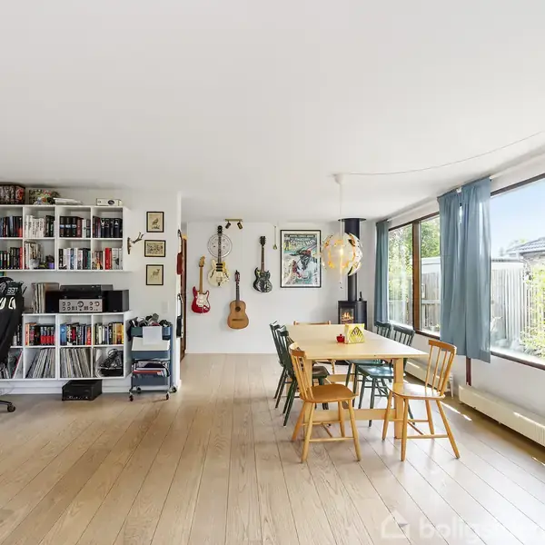 Stor stue med hvide og røde vægge. Sofa og billeder hænger på væggene. Døre åbner til en have med grønne buske og blomster. Ingen tekst udover "Boligsiden" markering.