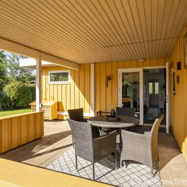 En overdækket terrasse med et rundt bord og fire flettede stole. Terrassen er af træ og malet gul, med en grøn have i baggrunden.