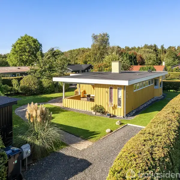 Et gult, moderne sommerhus med fladt tag står midt i en velplejet have med buske og grussti. Omkring huset ses grønne hække og et skur.
