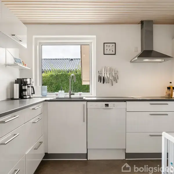 Et moderne køkken har hvide skabe, en kaffemaskine og knive hængende på væggen. Et vindue over vasken giver udsigt til en grøn hæk. En emhætte er placeret over komfuret.