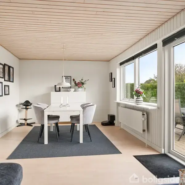 Et moderne spisestuebord står dækket af hvide stole og blomsterdekorationer. Rummet har store vinduer til en terrasse, træloft, billeder på væggen og et enkelt funktionelt design.