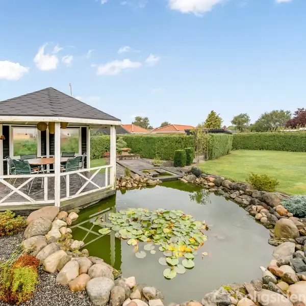 Pavillon med bord og stole står ved siden af en dam med åkander, omgivet af en græsplæne og buske i en velplejet have.