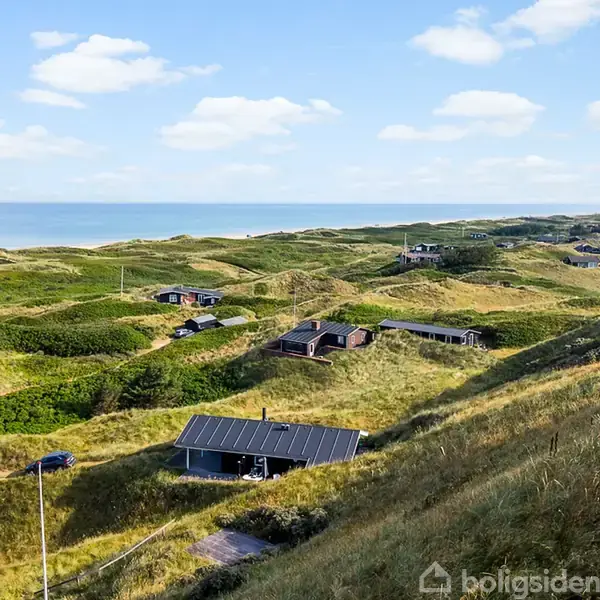 Stue med træmøbler og grå sofa i vinkel; en fladskærm står på et tv-bord. Store vinduer viser udsigt over bakket landskab, belysningen er lysekrone og væglampe.