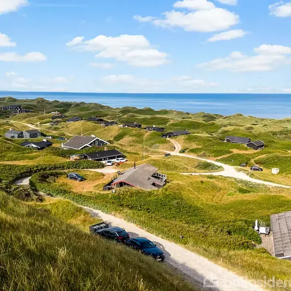 Sommerhuse ligger spredt i grønne klitter; nogle med parkerede biler ved siden af. I baggrunden ses havet under en let skyet, blå himmel.