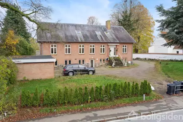 En to-etagers murstensbygning med skråt tag står i en grøn have. En sort bil er parkeret foran, nær en lille garagebygning. Træer omkranser området.