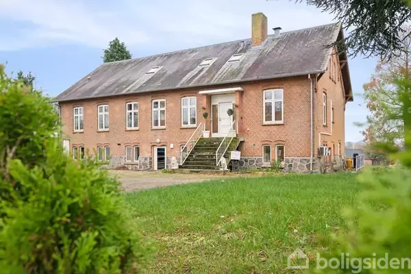Hus i røde mursten med tag af grå skifer, placeret i en grøn have. Flere vinduer og en bred trappe fører op til hoveddøren. Nogle træer omkranser bygningen.