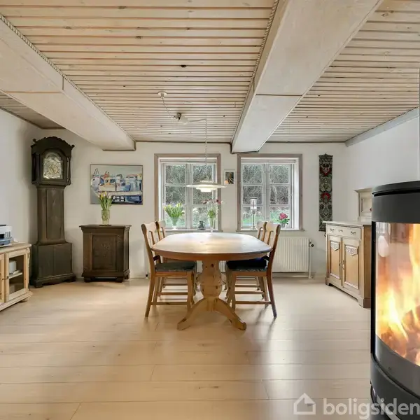 Brændeovn med ild i en hyggelig stue, plads til et spisebord og stole. Vinduer lader naturligt lys ind, omgivet af træmøbler og dekorationer på væggene.