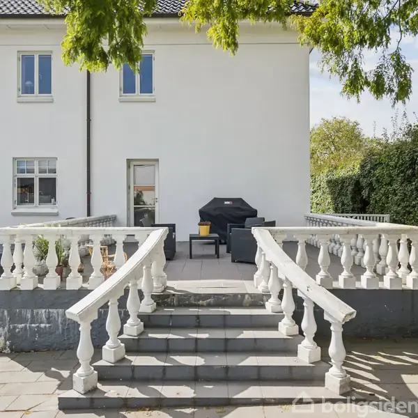 En terrasse med hvide balustre og trappe fører op til et sæt sorte havemøbler ved en hvid bygning. Omgivet af buske og træer under en blå himmel.