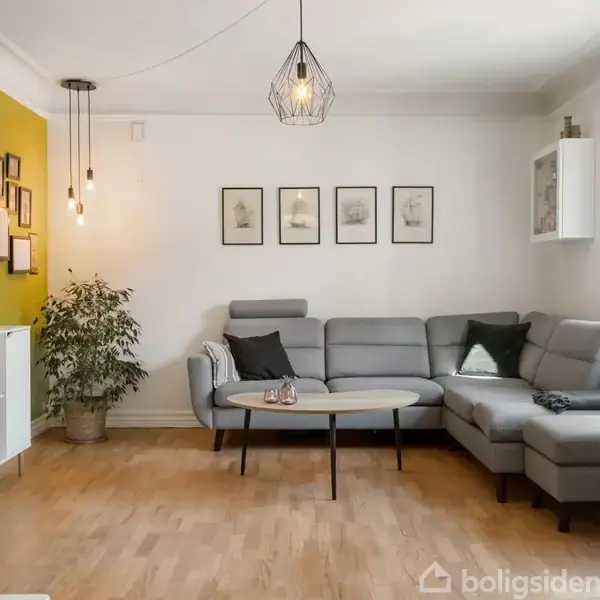 Grå sofa står i en lys stue med trægulv, flankeret af bord og puder. En grøn plante og vægpynt pryder rummet, som oplyses af moderne pendellamper.