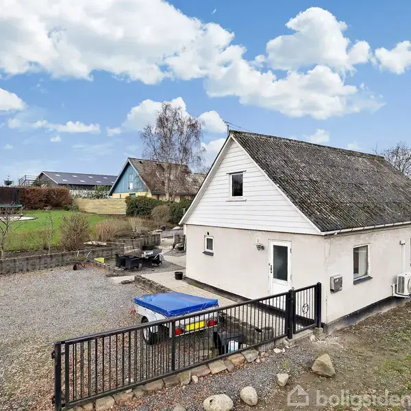 En hvid bygning med sadeltag står på en grusplads med en trailer udenfor. Bygningen er omgivet af en have med grønne græsarealer og andre huse i baggrunden.