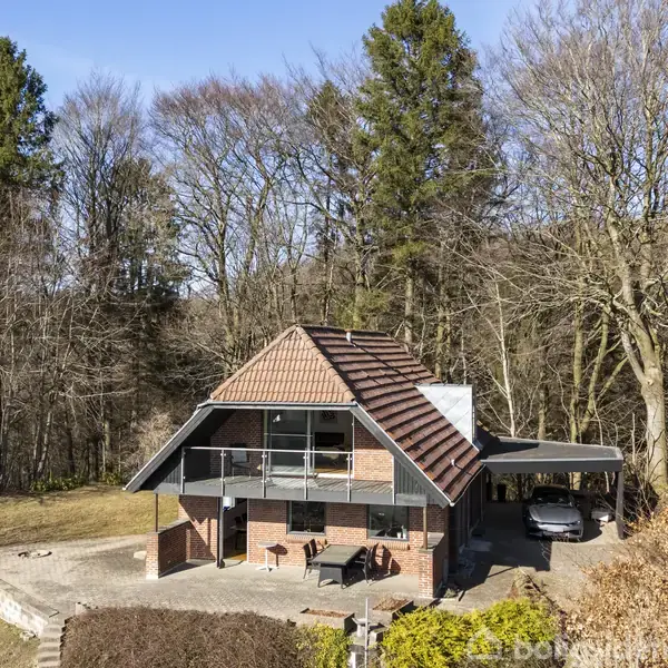 Hus af mursten med balkon står ved skoven. Der er en terrasse foran og en carport på siden. Omgivet af høje træer og buske.