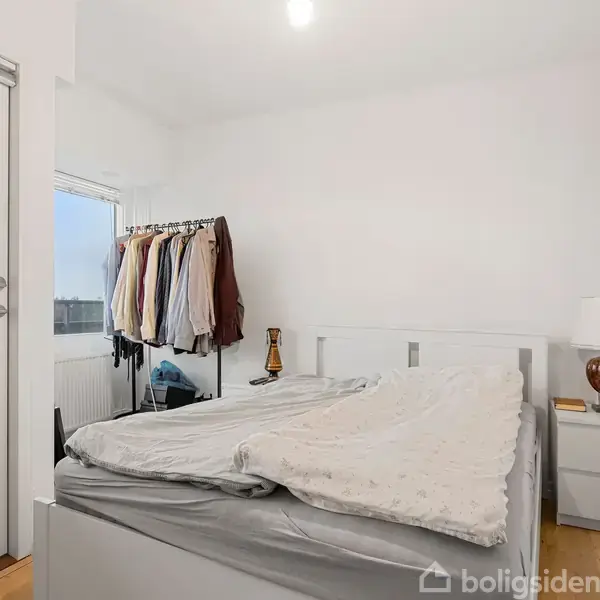 Seng placeret i et lyst soveværelse med tøjstativ ved siden af. Et natbord med en lampe står ved siden af sengen. En glasdør fører til en balkon.