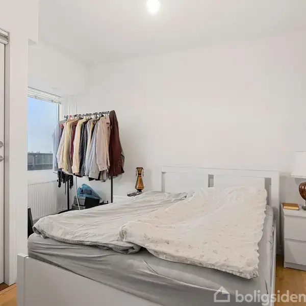Seng står opredt, omgivet af hvid væg. Tøj hænger på stang ved dør til altan. Natbord med lampe og bog ved siden af.