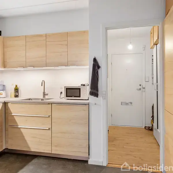 Køkken med lyse træskabe og sort gulv. En mikroovn og køkkenredskaber står ved vasken. En dør fører til en lille gang med trægulv og hvid dør.