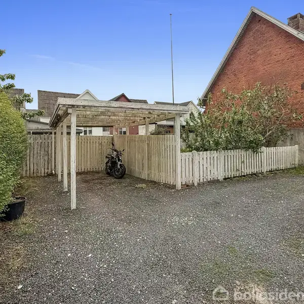 En motorcykel står parkeret under et træskur. Skuret er omgivet af et lavt hvidt stakit. Til højre ses en rød murstensbygning. Baggrunden viser flere hustage.