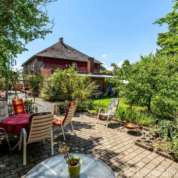 En terrasse med havemøbler står på brosten i en frodig have. I baggrunden ses et rødt hus omkranset af grønne planter, buske og træer, der skaber en hyggelig atmosfære.