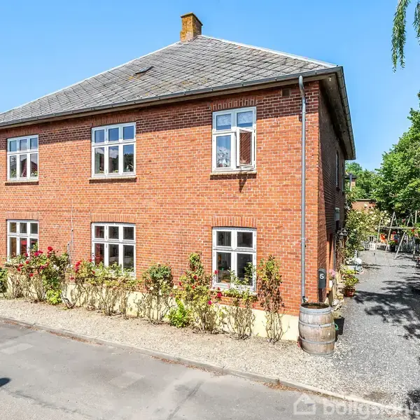 En rød murstensbygning med to etager står stille. Omkring den ses buske og blomster foran et grusbelagt gangareal. En blå himmel og grønne træer omgiver huset.