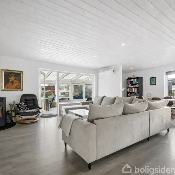 Et rundt spisebord med brune stole står i en moderne stue. En sofa vender mod et tv-modul. Bagerst anes et arbejdsområde med skrivebord og vinduer, der lyser rummet op.