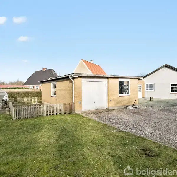 En lille garagebygning står stille på en grusplads i en have med grøn græsplæne; i baggrunden ses et drivhus og nabohuse under en blå himmel.