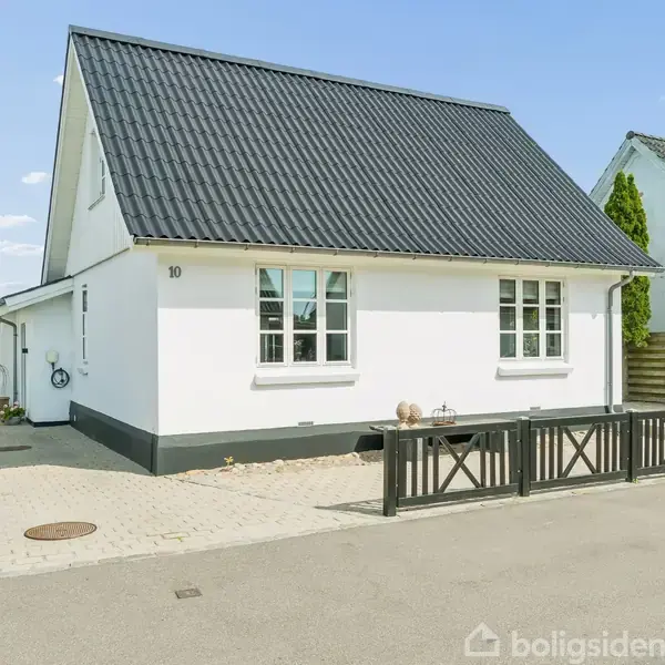 Et hvidt hus med sort tag står ved en vej. Huset har hvide vinduer, en lav sort stakit foran og buske i baggrunden. Nummeret "10" er på facaden.