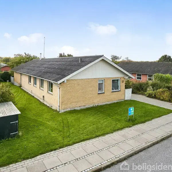 En gul murstensbungalow står stille i et grønt område med velplejet græsplæne, omgivet af en flisegang og en mindre havelåge ved vejen.