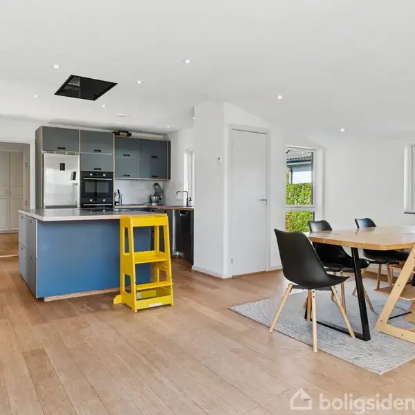 Køkkenalrum med spisebord, seks stole og et blåt køkken med ovn samt køleskab. Gul stol står ved køkkenøen. Store vinduer giver lys, og grønne buske ses udenfor.