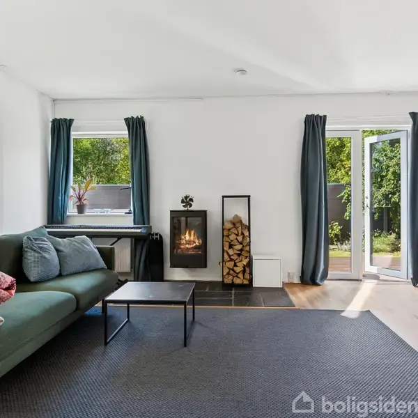 Sofa står i stue ved siden af en sort bord, mens en brændeovn brænder ved siden af et vindue. Døren åbner ud til en have med grønne buske.