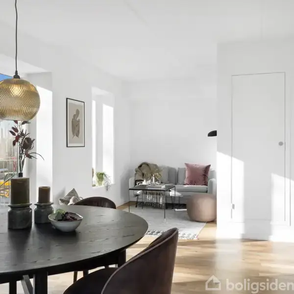 Rundt spisebord med mørke stole står i en lys stue. En moderne lampe hænger over bordet. Baggrunden viser en grå sofa med puder og glasvægge, som giver naturligt lys.