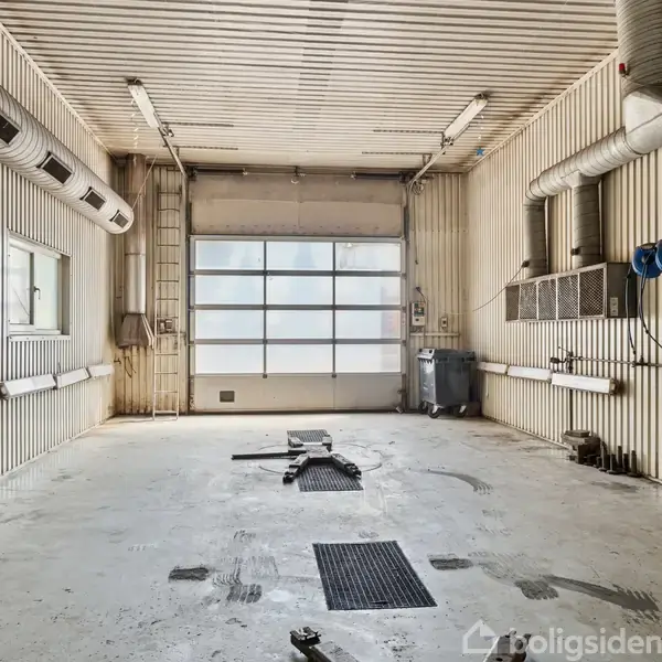 En tom garage med hvidmalede vægge og loft, ventilationsrør, og et stort glasporte. Mørke rektangulære paneler er installeret i gulvet. Værktøj og slanger hænger på højre væg.