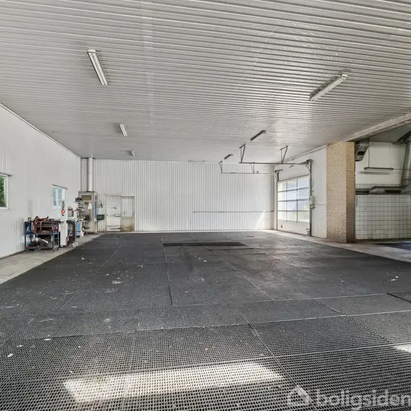 Et rummeligt, tomt værksted med sort gulv, hvide vægge og et loft med lysstofrør. En port til venstre indikerer ind- og udgangsveje. Ingen tekst at transkribere.