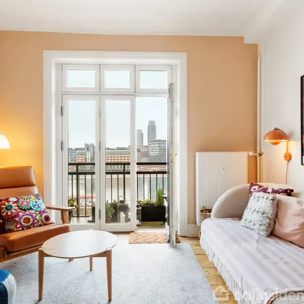 Stue med lænestol og sofa er arrangeret omkring et lille rundt bord. Stort vindue åbner ud til en altan med udsigt over byen. Plakater hænger på væggen.