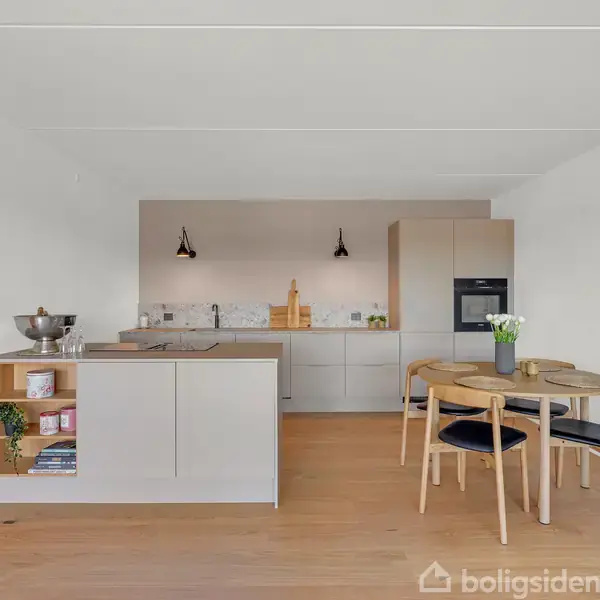 Et moderne køkken med gråt interiør; en køkkenø står i centrum med frugtskål og planter, omgivet af lyse træstole og bord; baggrunden har indbyggede skabe og ovn.