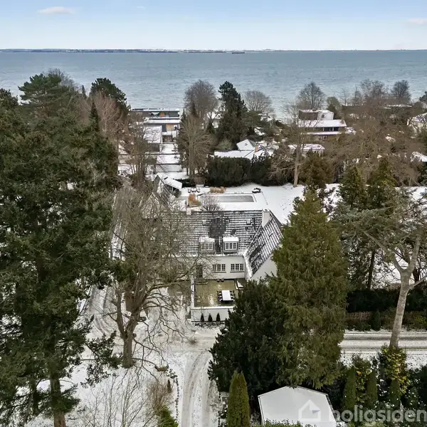Et hus omgivet af sneklædte træer og haver tæt ved en kystlinje. I baggrunden ses en stor vandmasse og andre ejendomme, der også er dækket af sne.