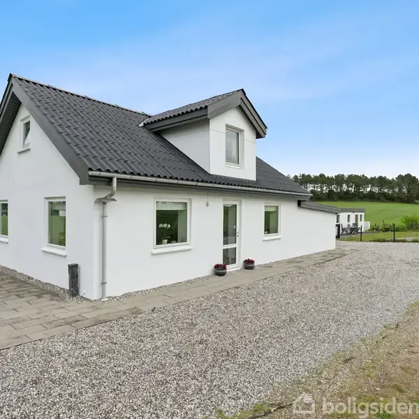 Hvidt hus med sort tag står stille på grusvej. Omkringliggende marker og en trælinje i baggrunden under en klar blå himmel.