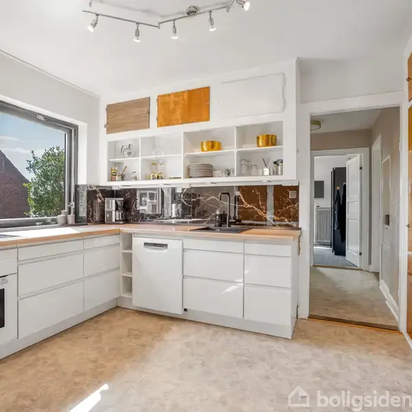 Et moderne køkken med hvide skabe og indbyggede apparater står i et lyst rum. En stor vindue lader naturligt lys ind. Korridor i baggrunden fører til andre rum.