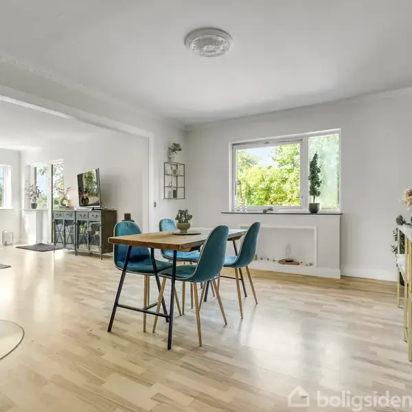 Et spisebord med blå stole står på et lyst trægulv. Vinduet bagved giver udsigt til træer. Der er en sort brændeovn til venstre i et moderne, lyst rum.
