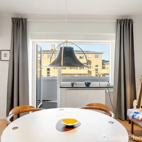 Rundt spisebord med en gul skål i en lys stue. Stuen har et vindue med grå gardiner, en lampe hænger over bordet, og der er en guitar ved en farverig pude.