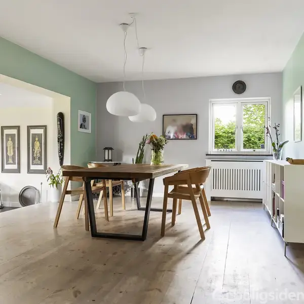 Et spisebord med træstole står i midten af en lys stue. Omkring bordet er der en grøn væg, vægkunst, en radiator under et vindue, og en åben passage til en anden del af huset.
