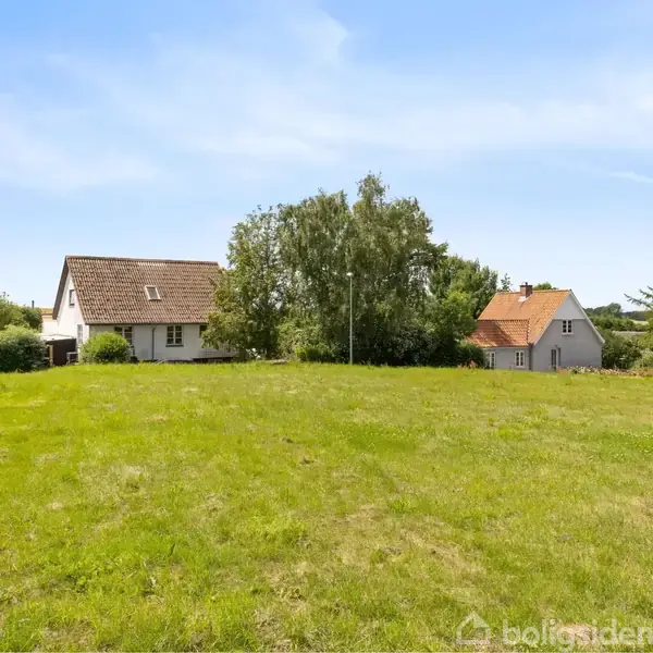 To huse står i en grøn have med græsplæne; et hvidt hus til venstre og et gråt hus til højre. Blå himmel med lette skyer danner baggrunden.