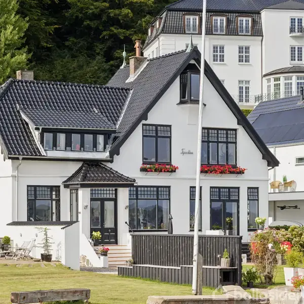 En hvid villa med sort tag, omgivet af en grøn have. Foran huset står en flagstang med Dannebrog. Huset har blomsterkasser med røde blomster under vinduerne.