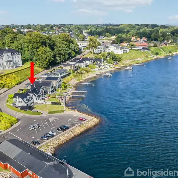 Hus markeret med rød pil. Omgivet af andre bygninger langs kysten ved en fjord. Bilparkering foran huset. Skoven i baggrunden.