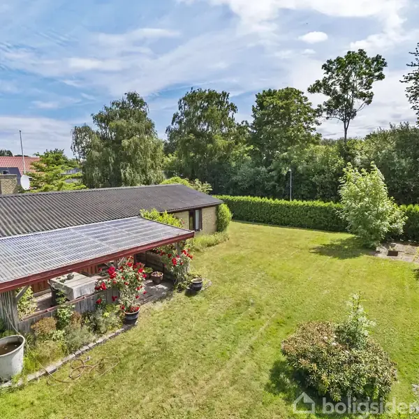 Hus med skråtag står i en velplejet have med grøn græsplæne, buske og træer. I baggrunden er der en tæt hæk og flere høje træer under en blå himmel.